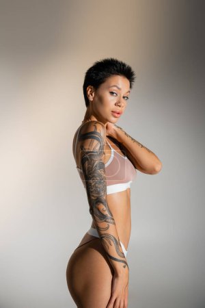 brunette woman with perfect tattooed body touching neck while posing in underwear on grey background