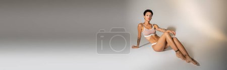 full length of fit woman with tattooed body posing in lingerie on grey background, banner