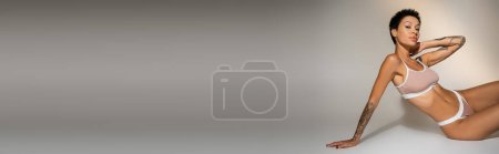 Photo for Brunette woman in underwear touching neck and looking at camera while sitting on grey background, banner - Royalty Free Image