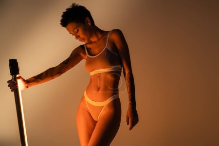 mujer morena tatuada con cuerpo delgado sosteniendo lámpara fluorescente mientras posa en lencería sobre fondo beige