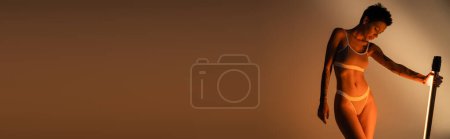 Téléchargez les photos : Femme tatouée en lingerie debout avec lampe fluorescente lumineuse sur fond sombre, bannière - en image libre de droit