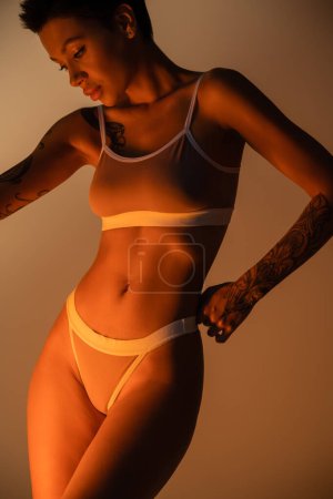 sinnliche brünette Frau mit perfektem Körper posiert in Dessous auf beigem Hintergrund mit Licht