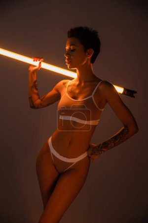 mujer tatuada en ropa interior sosteniendo lámpara fluorescente mientras posa con la mano en la cadera sobre fondo oscuro