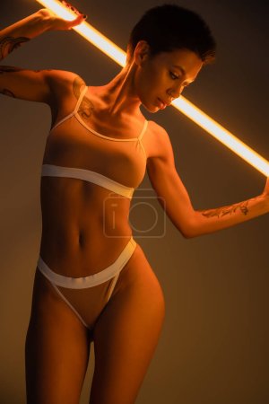 brunette woman with perfect tattooed body wearing underwear and posing with fluorescent lamp on dark background