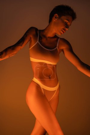 seductive tattooed woman in underwear posing in light on dark background