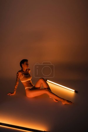 volle Länge der schlanken tätowierten Frau in Dessous, die neben lebendigen Leuchtstofflampen auf dunklem Hintergrund sitzt