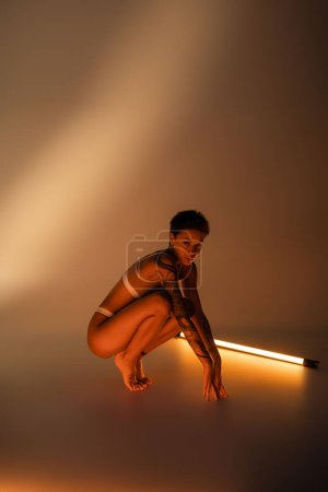 full length of tattooed woman in underwear sitting on haunches near fluorescent lamp on beige background