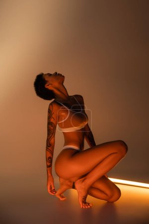 Téléchargez les photos : Side view of fit woman with perfect tattooed body posing in underwear near fluorescent lamp on beige background - en image libre de droit