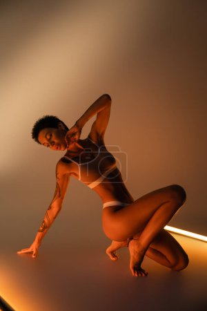 full length of seductive woman with tattooed and perfect body posing in underwear near fluorescent lamps on beige background