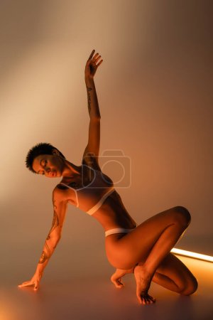 longitud completa de la sensual mujer joven en ropa interior posando con la mano levantada cerca de la lámpara fluorescente brillante sobre fondo beige