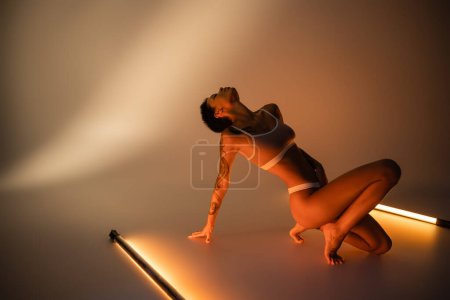 Foto de Longitud completa de morena mujer tatuada en ropa interior posando cerca de lámparas fluorescentes sobre fondo beige - Imagen libre de derechos