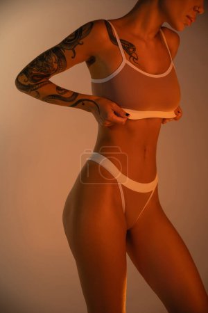 partial view of sexy woman with perfect tattooed body adjusting bra in light on beige background 