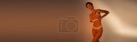 brunette woman with slim tattooed body adjusting bra and looking at camera on beige background, banner