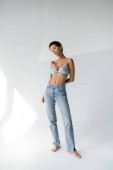 full length of tattooed barefoot woman in blue jeans and satin bra looking at camera while standing on grey background t-shirt #638597554
