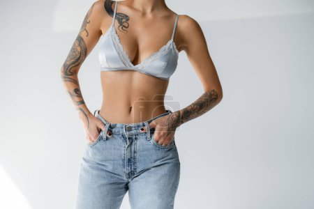 partial view of tattooed and slim woman in silk bralette holding hands in pockets of blue jeans on grey background magic mug #638597574