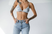 partial view of tattooed and slim woman in silk bralette holding hands in pockets of blue jeans on grey background Poster #638597574
