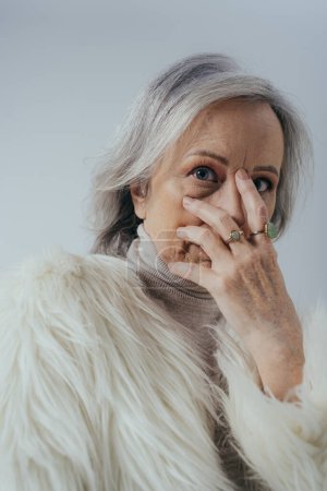 portrait of senior woman with rings on fingers looking at camera while covering face isolated on grey 