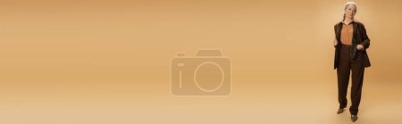 Téléchargez les photos : Longueur totale de la femme âgée en brun tenue formelle debout avec la main dans la poche sur fond beige, bannière - en image libre de droit