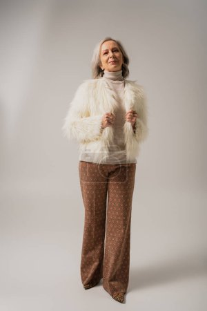 full length of pleased senior woman in faux fur jacket posing on grey background  