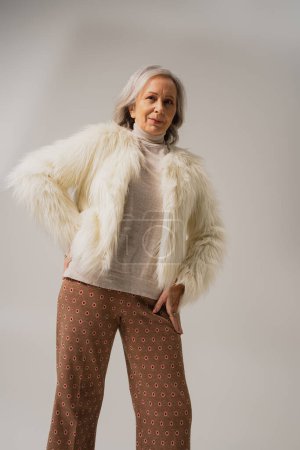 mujer mayor en chaqueta de piel sintética blanca posando con la mano en la cadera sobre fondo gris 