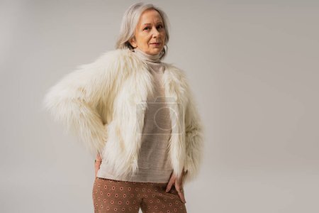 ältere Frau in weißer Kunstpelzjacke posiert mit der Hand an der Hüfte isoliert auf grau 