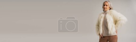 Téléchargez les photos : Femme âgée en fausse fourrure blanche posant à la main sur la hanche isolée sur gris, bannière - en image libre de droit