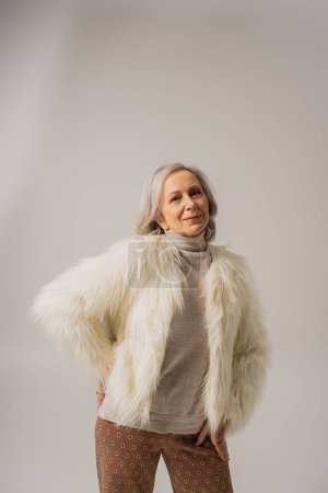mujer anciana complacida en chaqueta de piel sintética blanca posando con la mano en la cadera sobre gris 