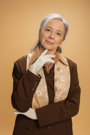 Téléchargez les photos : Portrait de femme âgée heureuse en gants blancs et costume marron regardant la caméra isolée sur beige - en image libre de droit