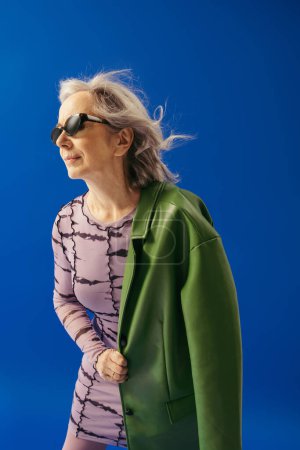 senior woman in dress and trendy sunglasses standing with green jacket isolated on blue 