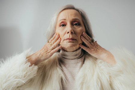 Téléchargez les photos : Portrait de femme âgée avec des anneaux sur les doigts regardant la caméra tout en touchant le visage sur fond gris - en image libre de droit