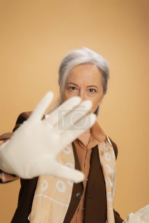 senior woman in white glove looking at camera while showing stop sign isolated on beige 