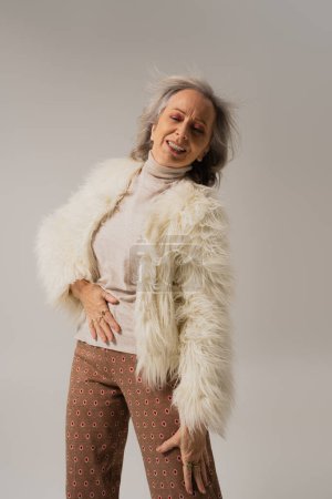 Emotionale Seniorin in Kunstpelzjacke posiert isoliert auf Grau 
