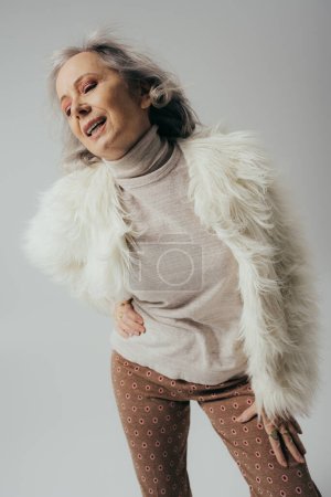 mujer mayor emocional en chaqueta de piel sintética posando con la mano en la cadera aislado en gris 