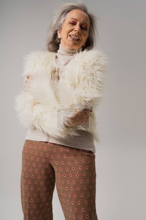 Emotionale Seniorin in Kunstpelzjacke posiert mit verschränkten Armen in Grau 