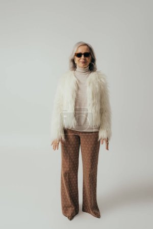 longitud completa de feliz anciana en chaqueta de piel sintética blanca y gafas de sol posando en gris 