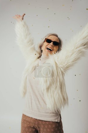 glückliche ältere Frau in weißer Kunstpelzjacke und Sonnenbrille, die die Hände neben fallendem Konfetti auf Grau hebt 