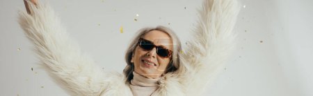 Photo for Cheerful elderly woman in white faux fur jacket and sunglasses raising hands near falling confetti on grey background, banner - Royalty Free Image
