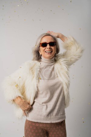 glückliche ältere Frau in weißer Kunstpelzjacke und Sonnenbrille lächelt neben fallendem Konfetti auf Grau 