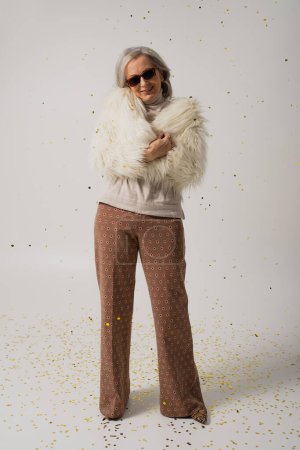longitud completa de alegre anciana en chaqueta de piel sintética blanca y gafas de sol de pie cerca de confeti caída sobre fondo gris 