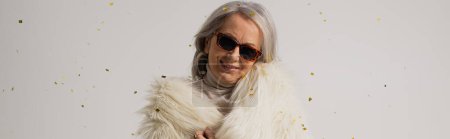 Téléchargez les photos : Pleased elderly woman in white faux fur jacket and sunglasses smiling near falling confetti on grey background, banner - en image libre de droit