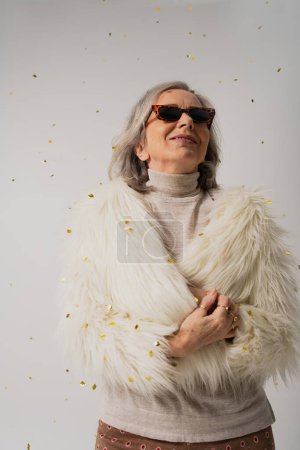 Téléchargez les photos : Heureux femme âgée en fausse fourrure blanche veste et lunettes de soleil souriant près de tomber confettis sur fond gris - en image libre de droit