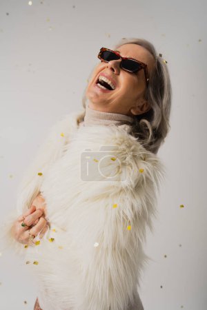 glückliche ältere Frau in weißer Kunstpelzjacke und trendiger Sonnenbrille lacht neben fallendem Konfetti auf Grau 