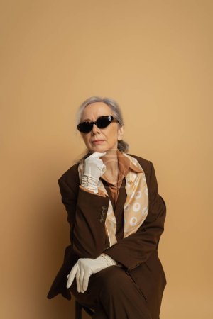 Nachdenkliche Seniorin in braunem Anzug und Sonnenbrille sitzt auf beigem Stuhl 