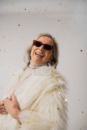 fröhliche ältere Frau in weißer Kunstpelzjacke und trendiger Sonnenbrille lacht neben fallendem Konfetti auf grauem Hintergrund 