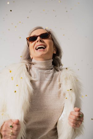 Photo for Positive elderly woman in white faux fur jacket and trendy sunglasses laughing near falling confetti on grey background - Royalty Free Image
