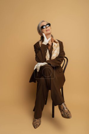 volle Länge der nachdenklichen Seniorin in brauner formeller Kleidung und Sonnenbrille sitzt auf einem Holzstuhl auf beige 