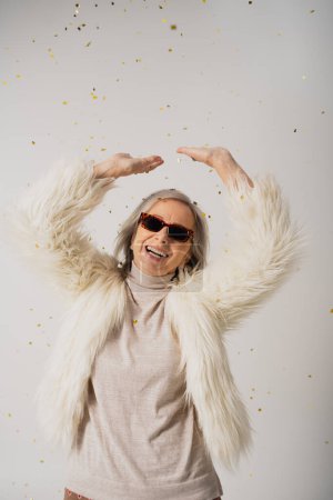 glückliche ältere Frau in weißer Kunstpelzjacke und Sonnenbrille, die mit erhobenen Händen neben fallendem Konfetti auf Grau steht 