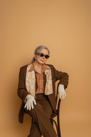 Foto de Mujer de edad avanzada en traje marrón y gafas de sol sentado en la silla y mirando a la cámara en beige - Imagen libre de derechos