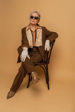 full length of elderly woman in brown suit and gloves sitting on chair and looking at camera on beige 