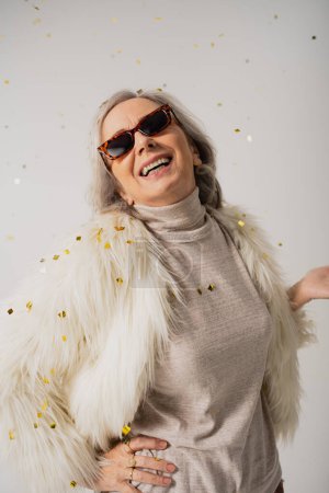 Foto de Mujer anciana complacida en chaqueta de piel sintética blanca y gafas de sol de moda sonriendo cerca de confeti caída sobre fondo gris - Imagen libre de derechos